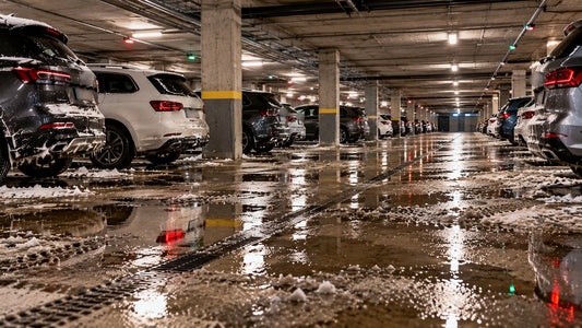Parkeringsanlegg under et boligkompleks med snødekte biler, smeltevann og slaps på betonggulvet og en dreneringsrenne.